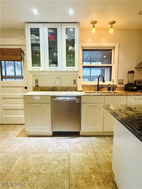 Kitchen featuring light stone countertops, stainless steel dishwasher, light tile patterned floors, white cabinets, and glass insert cabinets