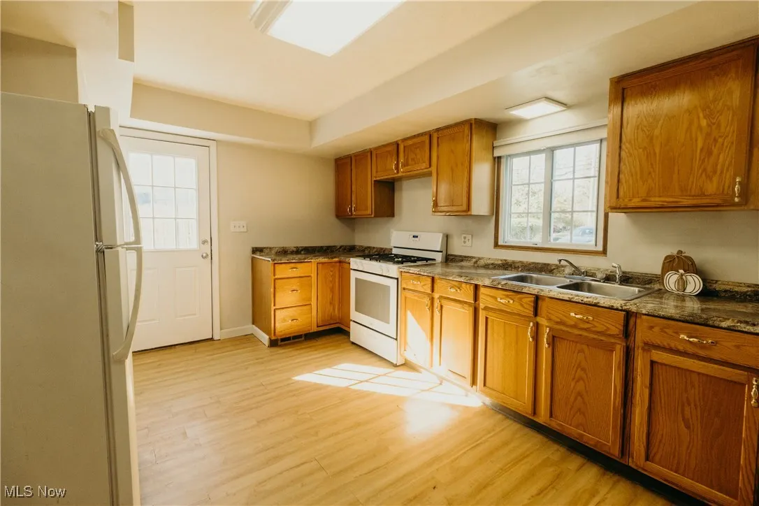 Kitchen featuring white appliances, brown cabinetry, light wood-style flooring, and dark countertops