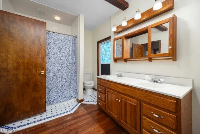 Full bathroom with double vanity, a shower with shower curtain, dark wood-type flooring, and beam ceiling