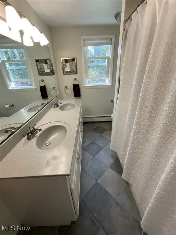Full bath with double vanity, a shower with shower curtain, a baseboard radiator, and dark tile patterned floors