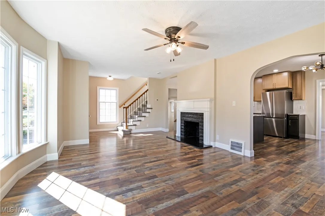 Unfurnished living room with arched walkways, stairs, a ceiling fan, a fireplace, and dark wood finished floors