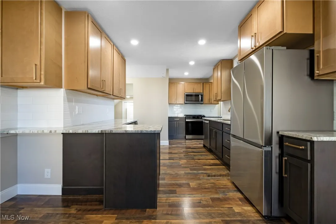 Kitchen with stainless steel appliances, light stone counters, decorative backsplash, dark wood-style floors, and a peninsula