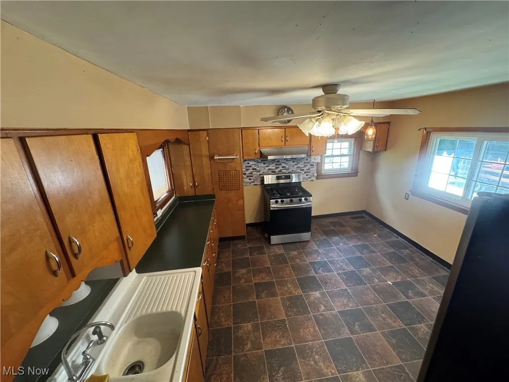 Kitchen featuring brown cabinets, stainless steel gas stove, decorative backsplash, stone finish flooring, and under cabinet range hood