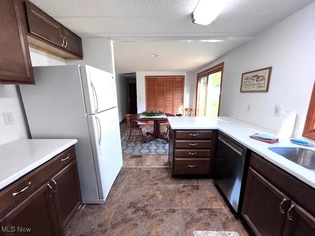 Kitchen featuring dark brown self close cabinetry, freestanding refrigerator, stainless steel dishwasher, and light Corian countertops. Tile flooring for easy clean ups.