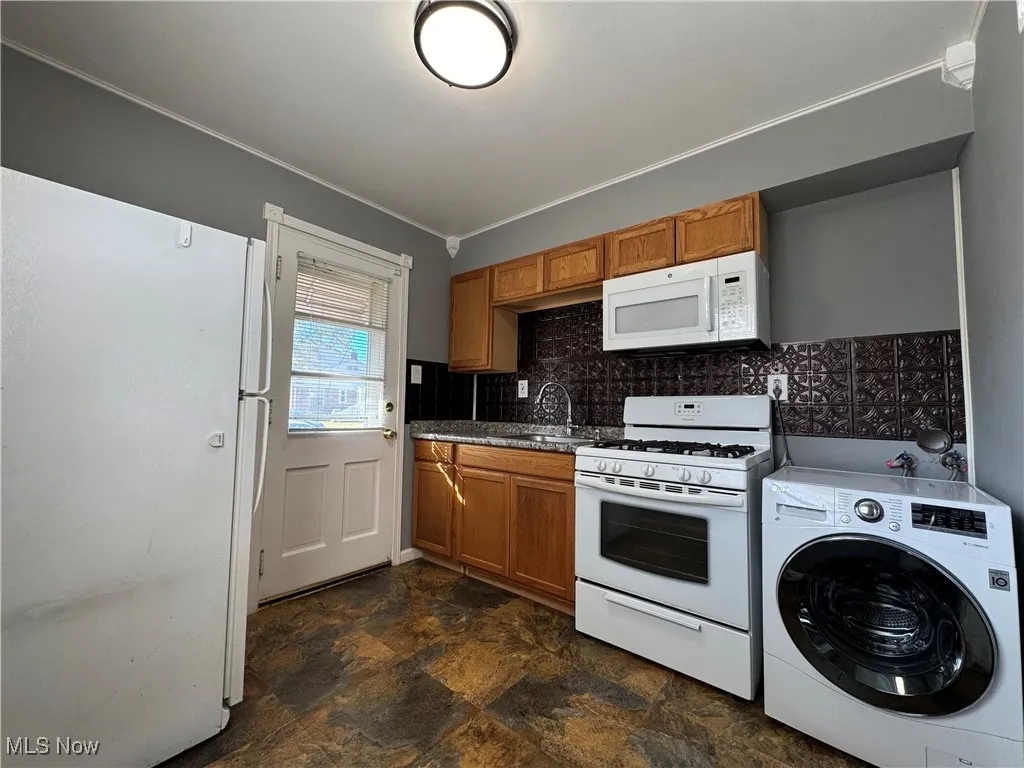 Kitchen featuring white appliances, washer / clothes dryer, stone finish floors, backsplash, and brown cabinets