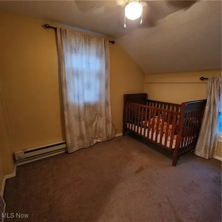 Carpeted bedroom with a textured ceiling, a ceiling fan, lofted ceiling, a nursery area, and a baseboard heating unit