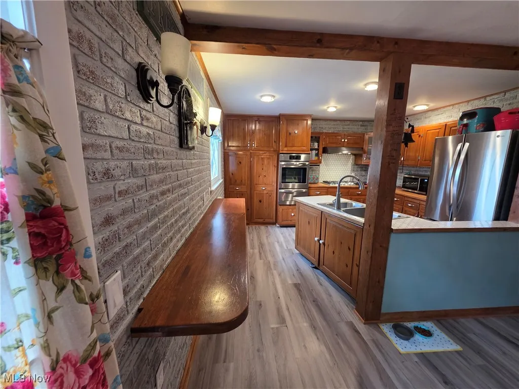 Kitchen featuring stainless steel appliances, brick wall, brown cabinetry, light wood-type flooring, and light countertops