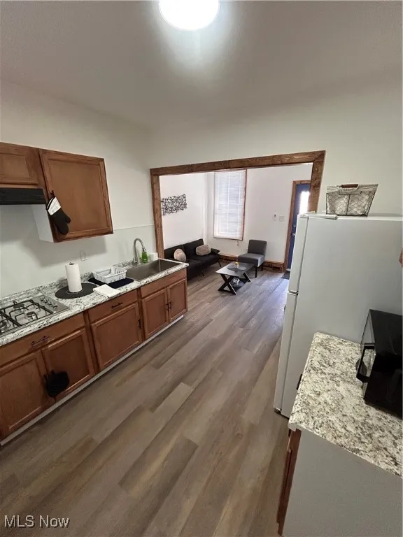 Kitchen featuring refrigerator, dark wood-type flooring, brown cabinetry, cooktop, and light stone counters