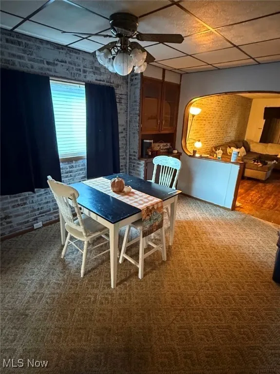 Carpeted dining area featuring a drop ceiling, brick wall, and ceiling fan