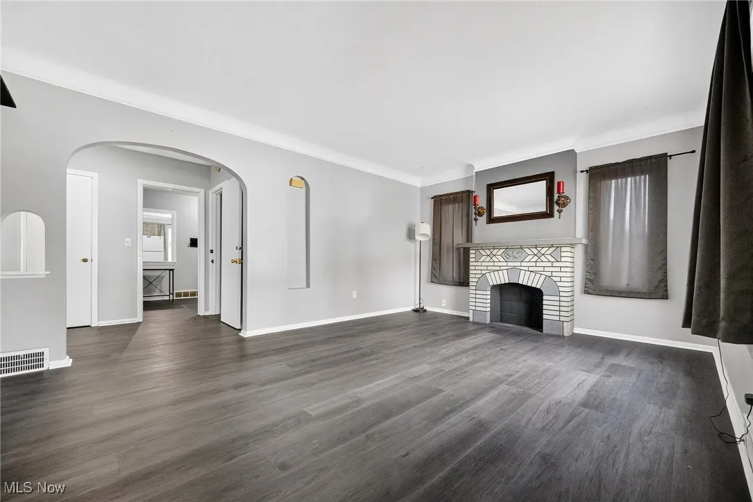 Unfurnished living room with dark wood-style floors, arched walkways, a brick fireplace, and crown molding