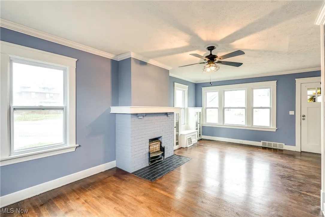 Unfurnished living room featuring crown molding, hardwood floors, a decorative fireplace, and ceiling fan.  Most windows have been replaced.