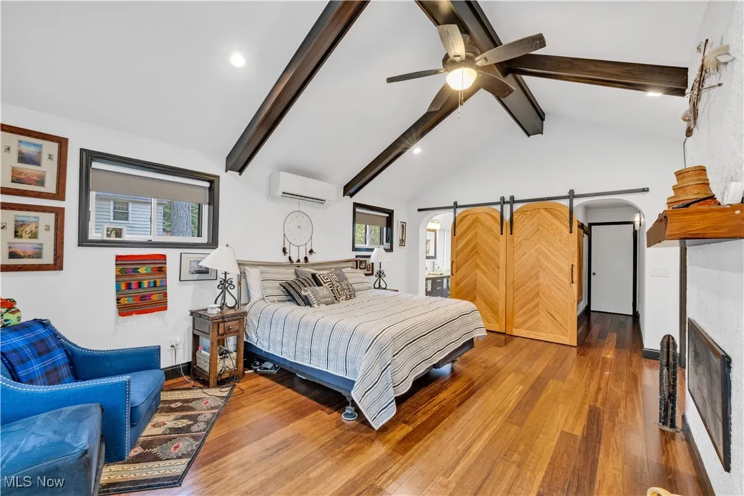 Bedroom with a barn door, wood finished floors, multiple windows, beamed ceiling, and connected bathroom