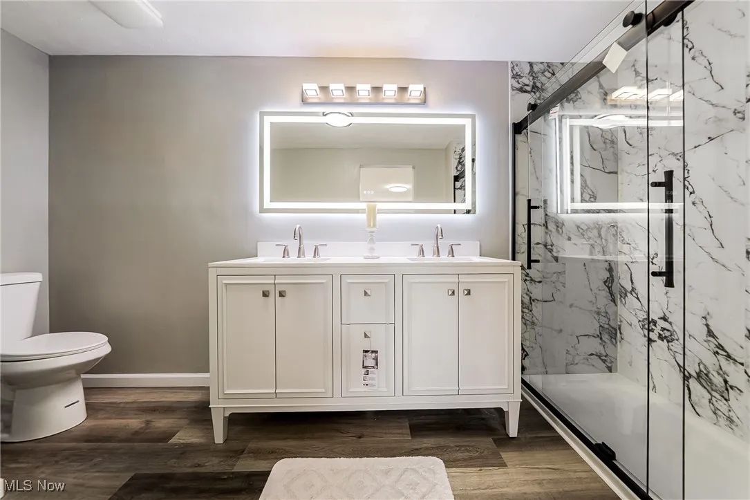Full bath featuring double vanity, a marble finish shower, and dark wood-type flooring