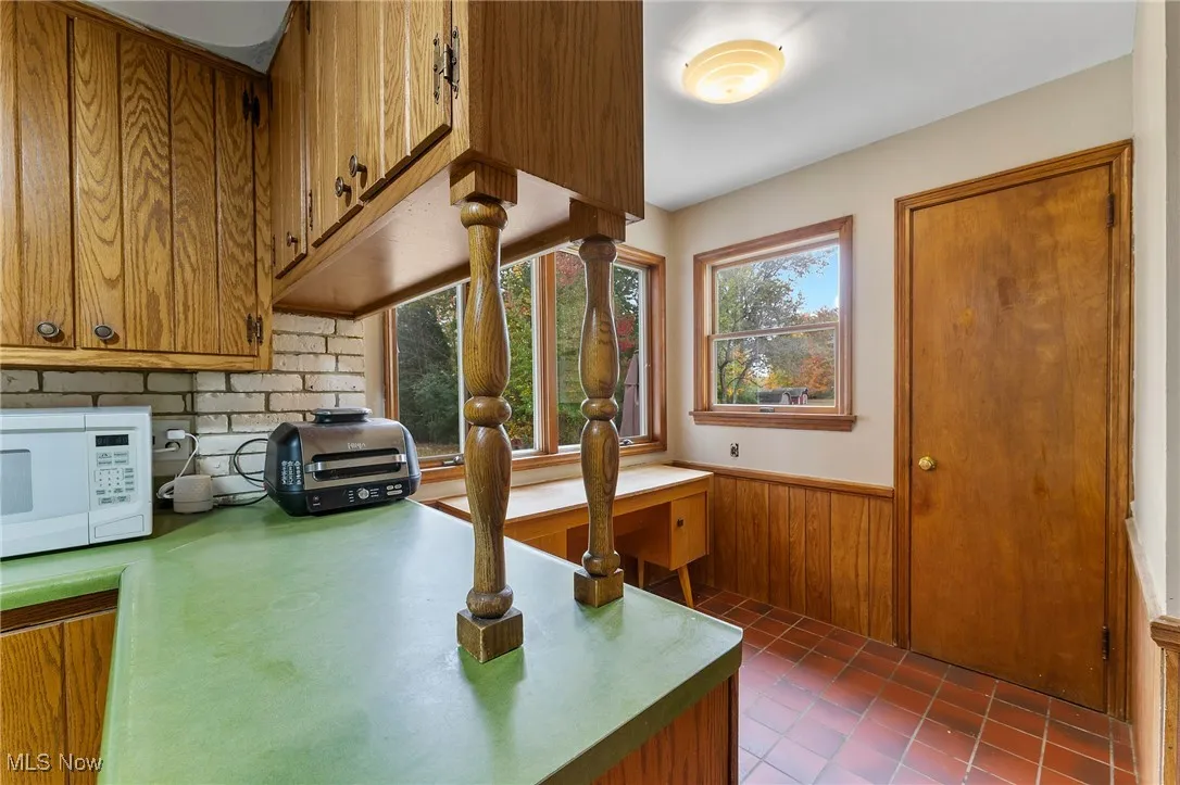 Kitchen featuring brown cabinets, wainscoting, white microwave, light countertops, and dark tile patterned floors