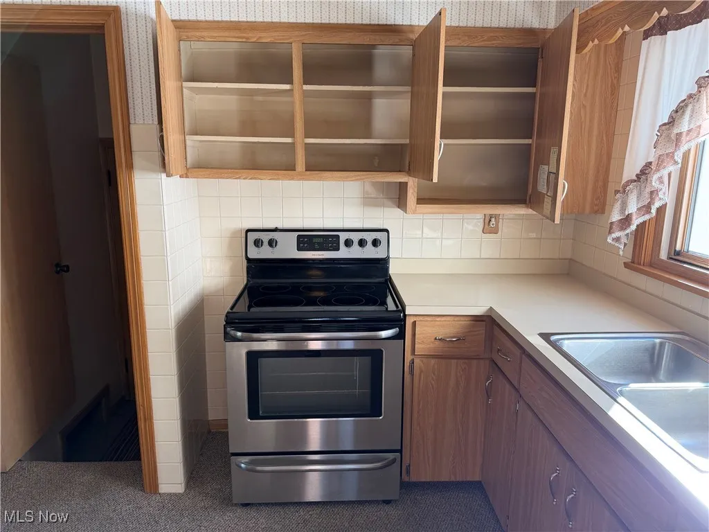 Kitchen with stainless steel electric stove, light countertops, brown cabinets, open shelves, and decorative backsplash
