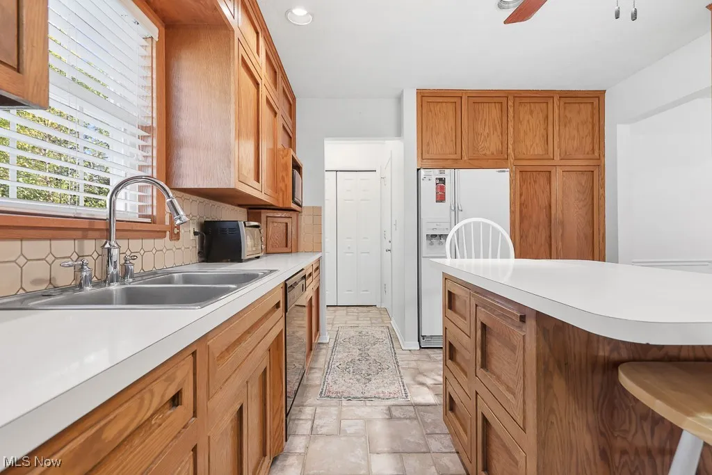 Kitchen with brown cabinets, white refrigerator with ice dispenser, light stone finish floors, light countertops, and tasteful backsplash