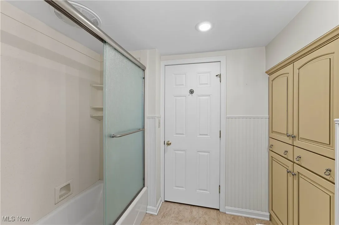 Bathroom with bath / shower combo with glass door, a wainscoted wall, light tile patterned floors, and recessed lighting