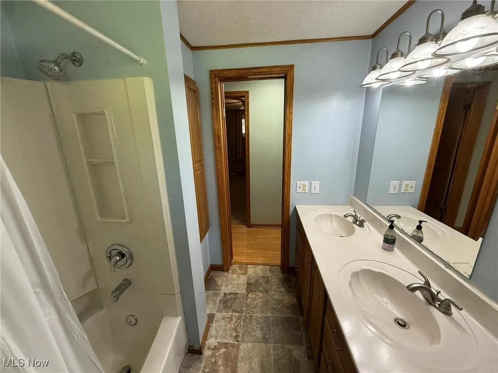 Full bath featuring crown molding, shower / bath combo with shower curtain, double vanity, stone finish floors, and a textured ceiling