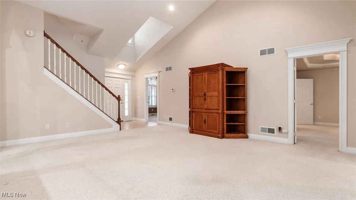 Unfurnished living room with high vaulted ceiling, light carpet, stairway, and a skylight