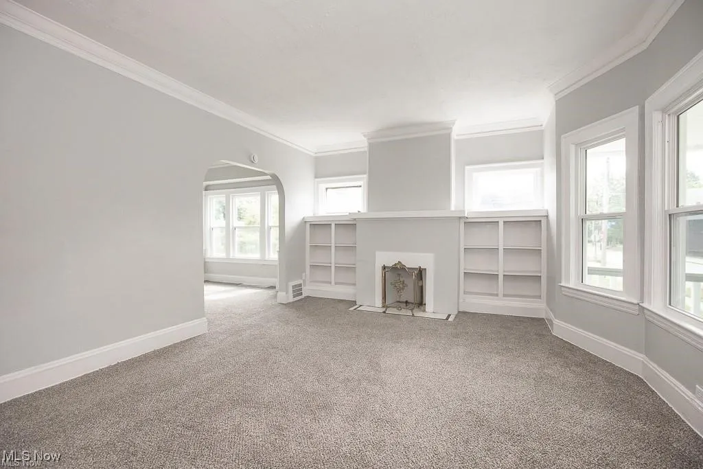Unfurnished living room featuring arched walkways, light colored carpet, and ornamental molding