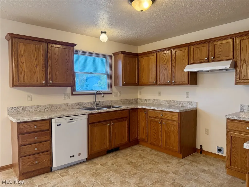 Kitchen with brown cabinets, white dishwasher, a textured ceiling, and under cabinet range hood