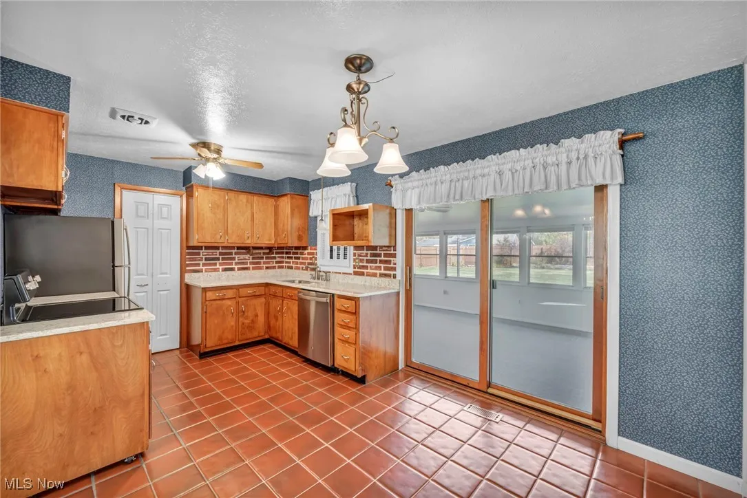 Kitchen with wallpapered walls, light tile patterned floors, stainless steel appliances, hanging light fixtures, and brown cabinetry