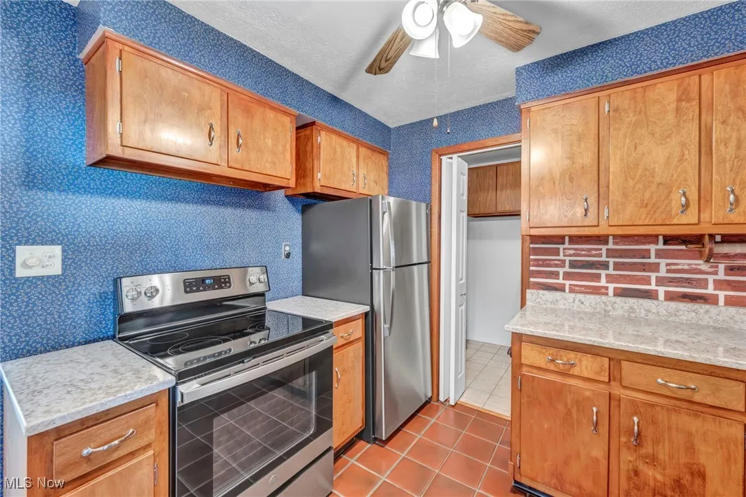 Kitchen with appliances with stainless steel finishes, wallpapered walls, light tile patterned flooring, ceiling fan, and light stone countertops