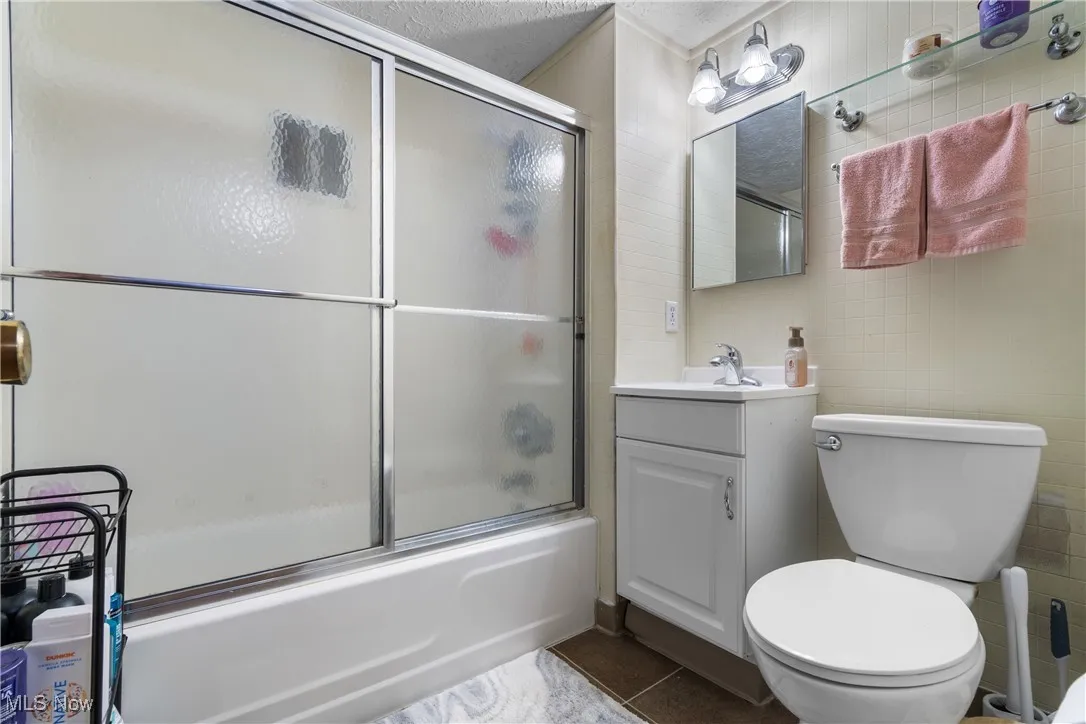 Bathroom featuring tile walls, dark tile patterned floors, vanity, bath / shower combo with glass door, and a textured ceiling
