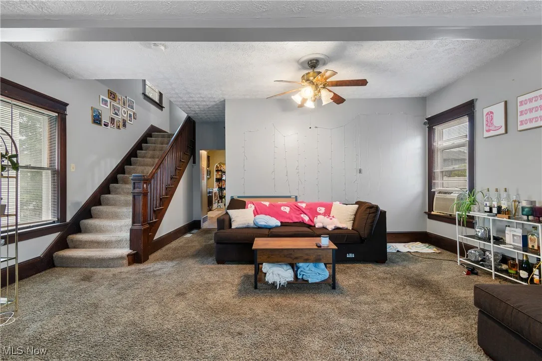 Carpeted living area featuring a textured ceiling, beamed ceiling, stairs, and ceiling fan