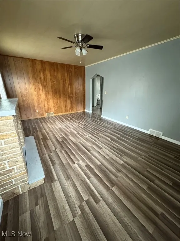 Unfurnished living room with arched walkways, wooden walls, dark wood-style flooring, crown molding, and ceiling fan