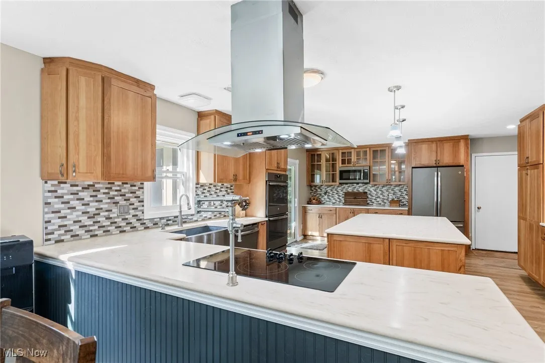 Kitchen featuring island range hood, tasteful backsplash, stainless steel appliances, glass insert cabinets, and light stone counters