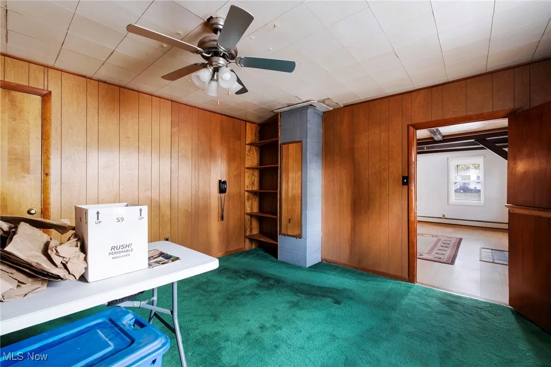 Unfurnished office with dark carpet, wood walls, a baseboard heating unit, and a ceiling fan
