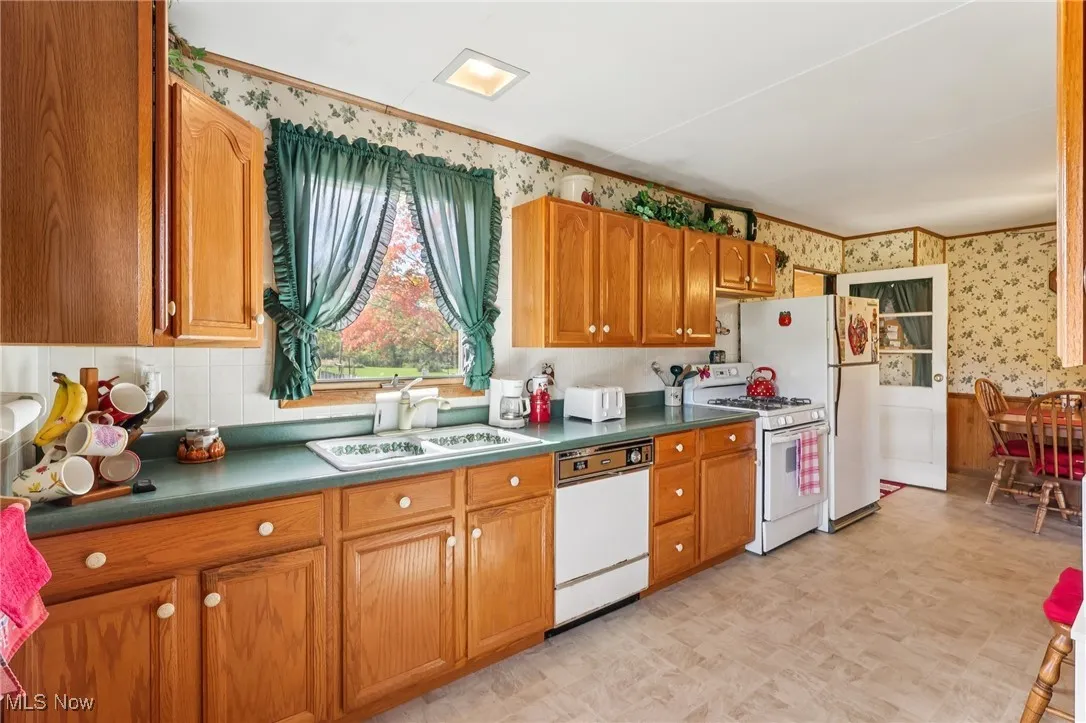 Kitchen with wallpapered walls, brown cabinets, white appliances, dark countertops, and light floors