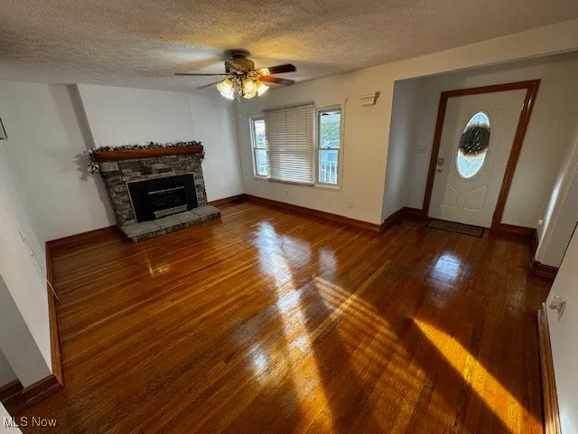Unfurnished living room with a textured ceiling, a ceiling fan, dark wood finished floors, and a stone fireplace