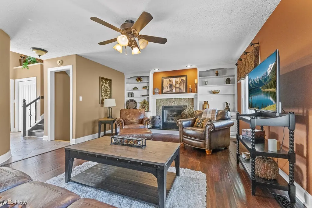 Living room with a tile fireplace, a textured ceiling, built in shelves, a ceiling fan, and dark wood-style flooring