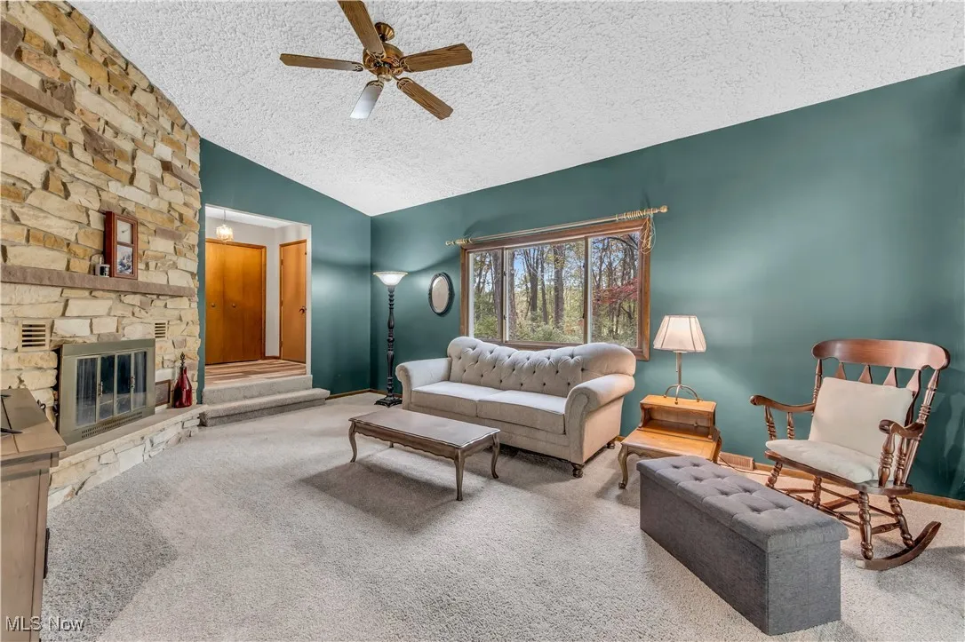 Carpeted living area featuring a textured ceiling, vaulted ceiling, a stone fireplace, and a ceiling fan