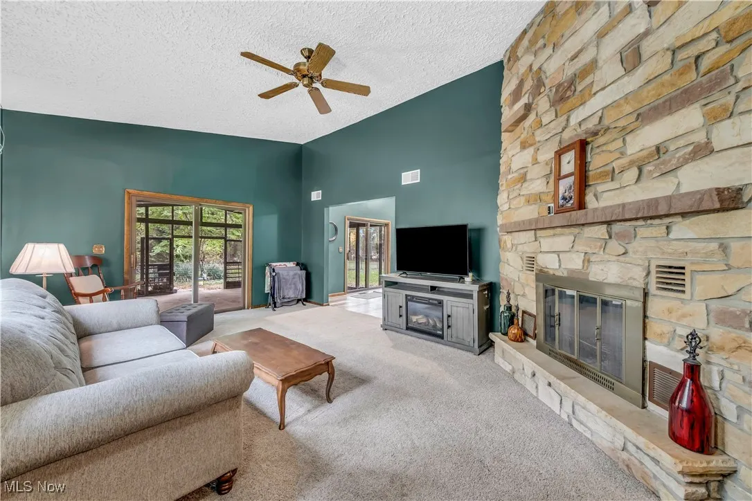 Living room featuring a stone fireplace, carpet flooring, a textured ceiling, a ceiling fan, and high vaulted ceiling