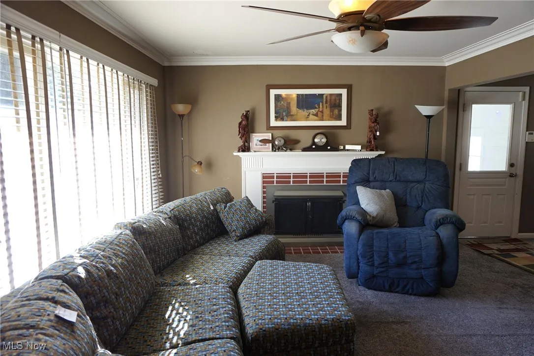 Living area featuring a glass covered fireplace, a ceiling fan, carpet flooring, and ornamental, crown molding.