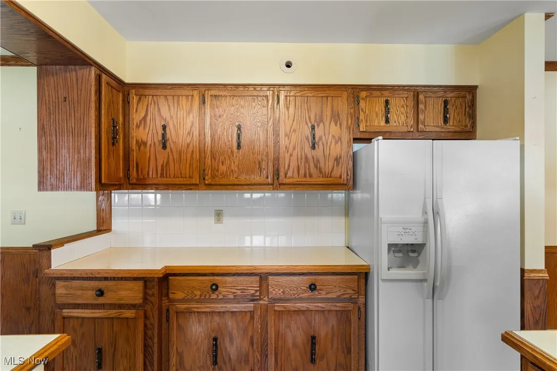 Kitchen featuring white fridge with ice dispenser, light countertops, brown cabinetry, wainscoting, and tasteful backsplash