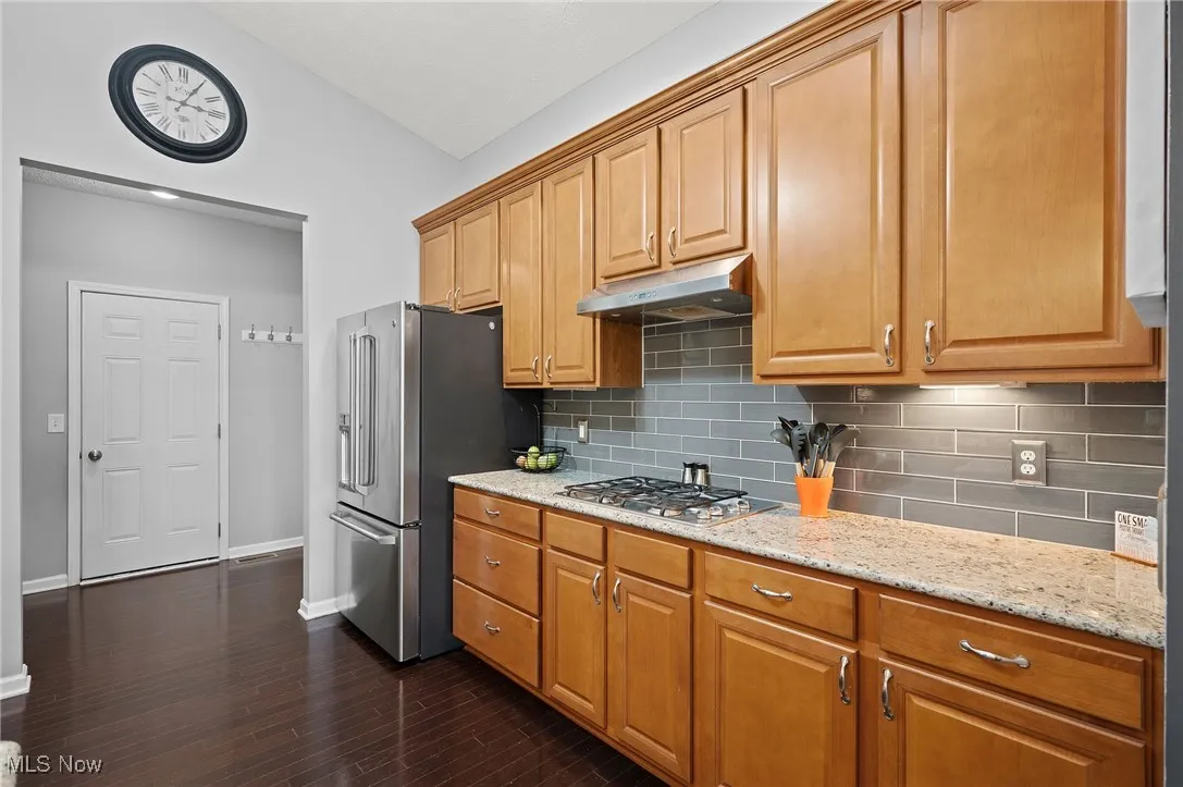 Kitchen with backsplash, light stone countertops, appliances with stainless steel finishes, brown cabinetry, and dark wood-type flooring