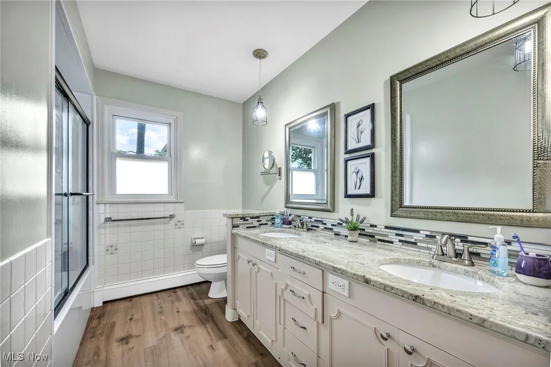 Bathroom with dark wood-type flooring, tile walls, double vanity, combined bath / shower with glass door, and wainscoting