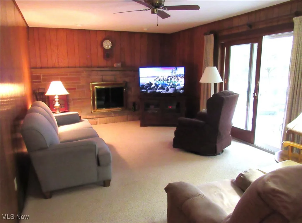 Carpeted living area with wooden walls, a fireplace, and ceiling fan