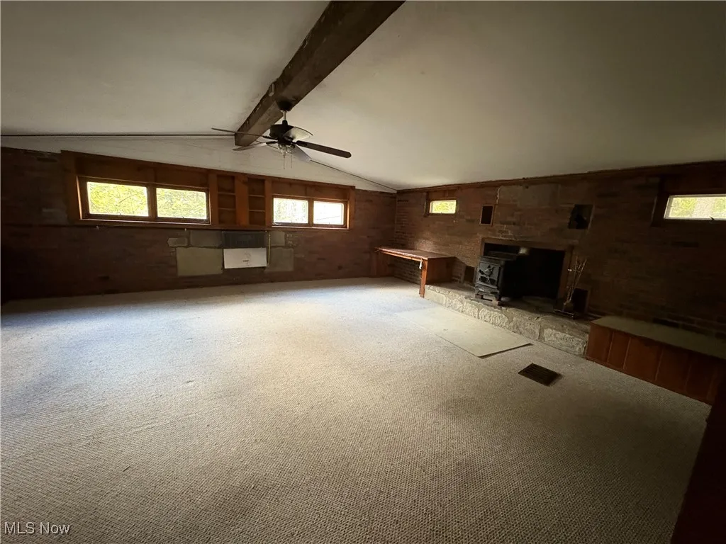 Unfurnished living room featuring a wood stove, carpet flooring, ceiling fan, heating unit, and brick wall