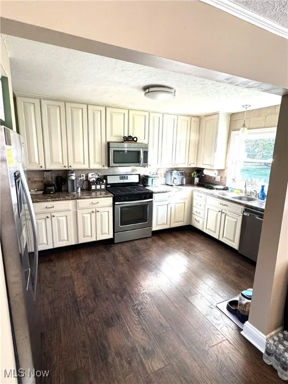 Kitchen with a textured ceiling, appliances with stainless steel finishes, dark wood-style flooring, cream cabinetry, and decorative light fixtures