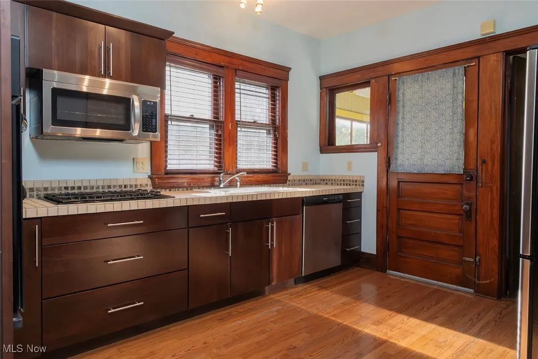 Kitchen with tile counters, dark brown wood cabinetry, stainless steel appliances, and light wood-type flooring