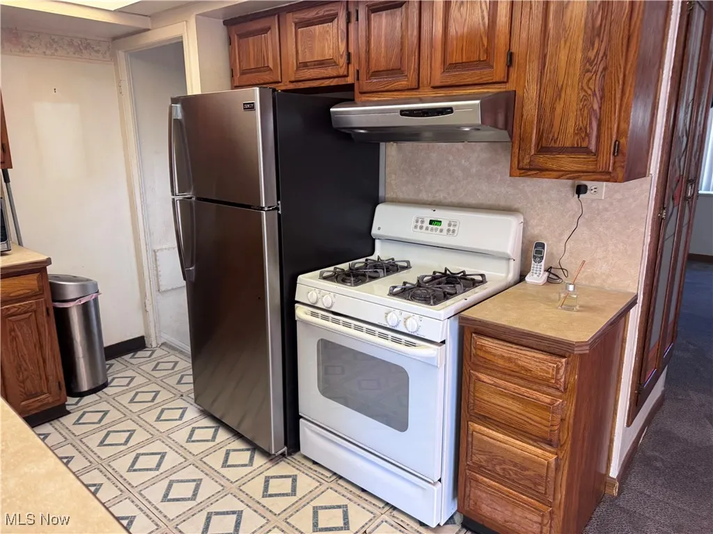 Kitchen with white range with gas cooktop, light countertops, brown cabinetry, range hood, and freestanding refrigerator