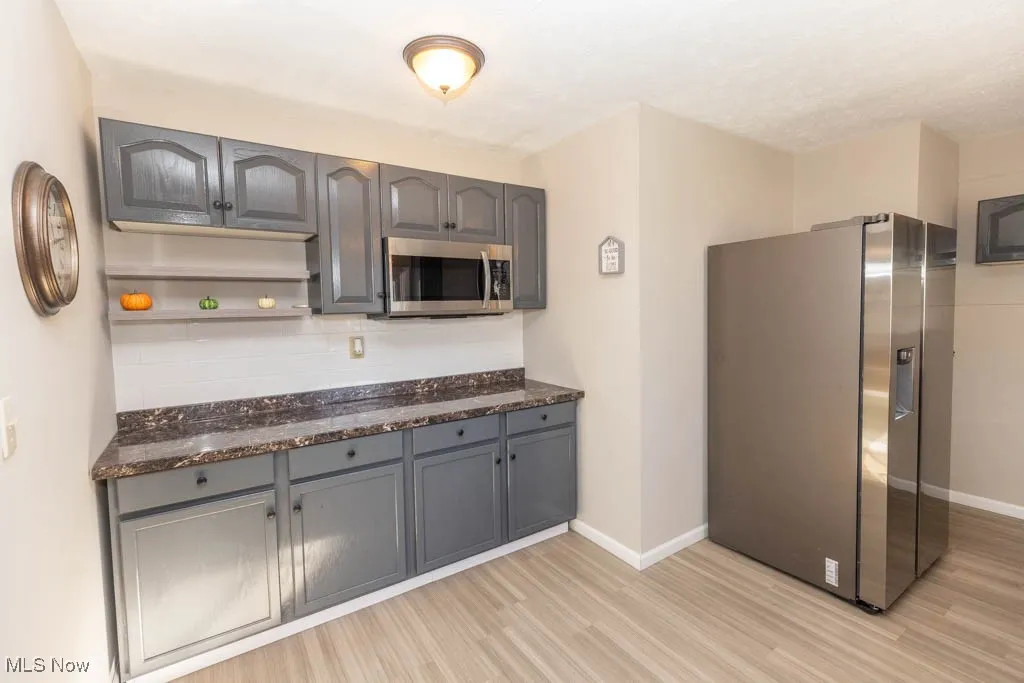 Kitchen with gray cabinetry, stainless steel appliances, light wood-type flooring, and dark stone countertops