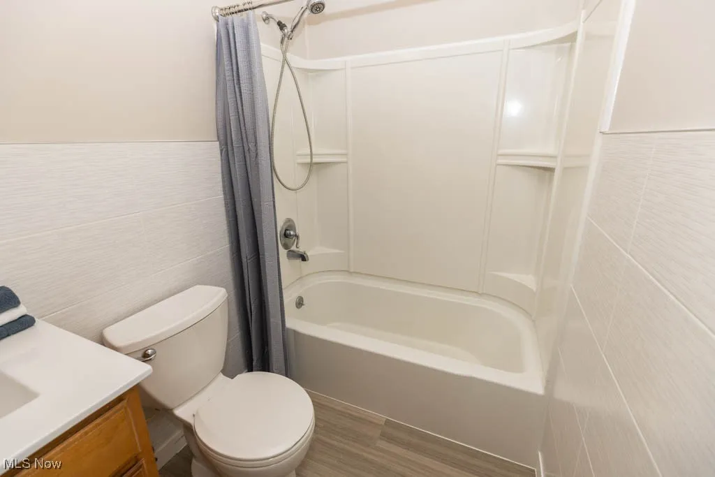 Full bath featuring tile walls, shower / bathtub combination with curtain, wood finished floors, wainscoting, and vanity