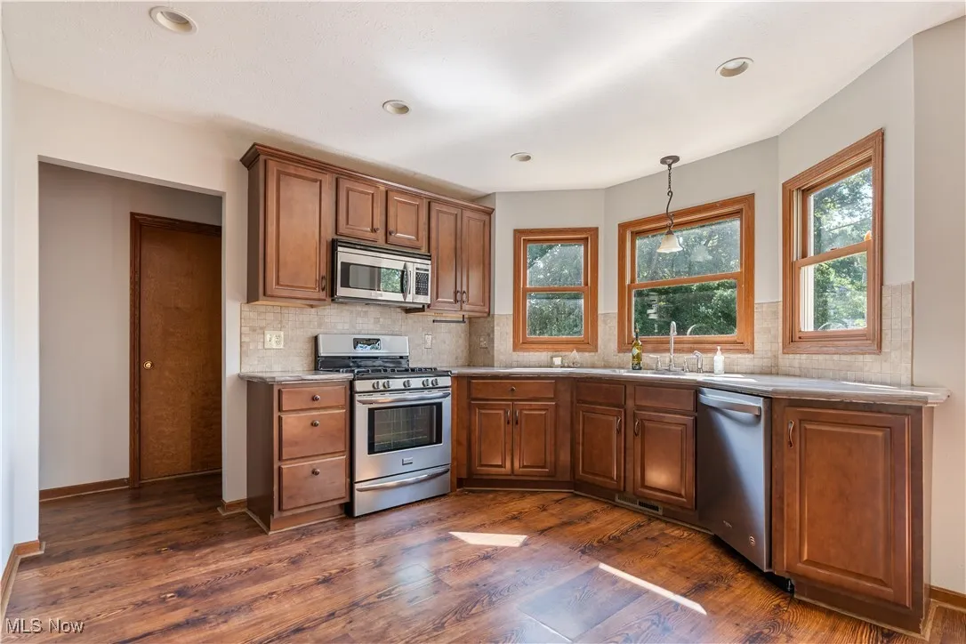 Kitchen with stainless steel appliances, hanging light fixtures, brown cabinetry, backsplash, and dark wood finished floors