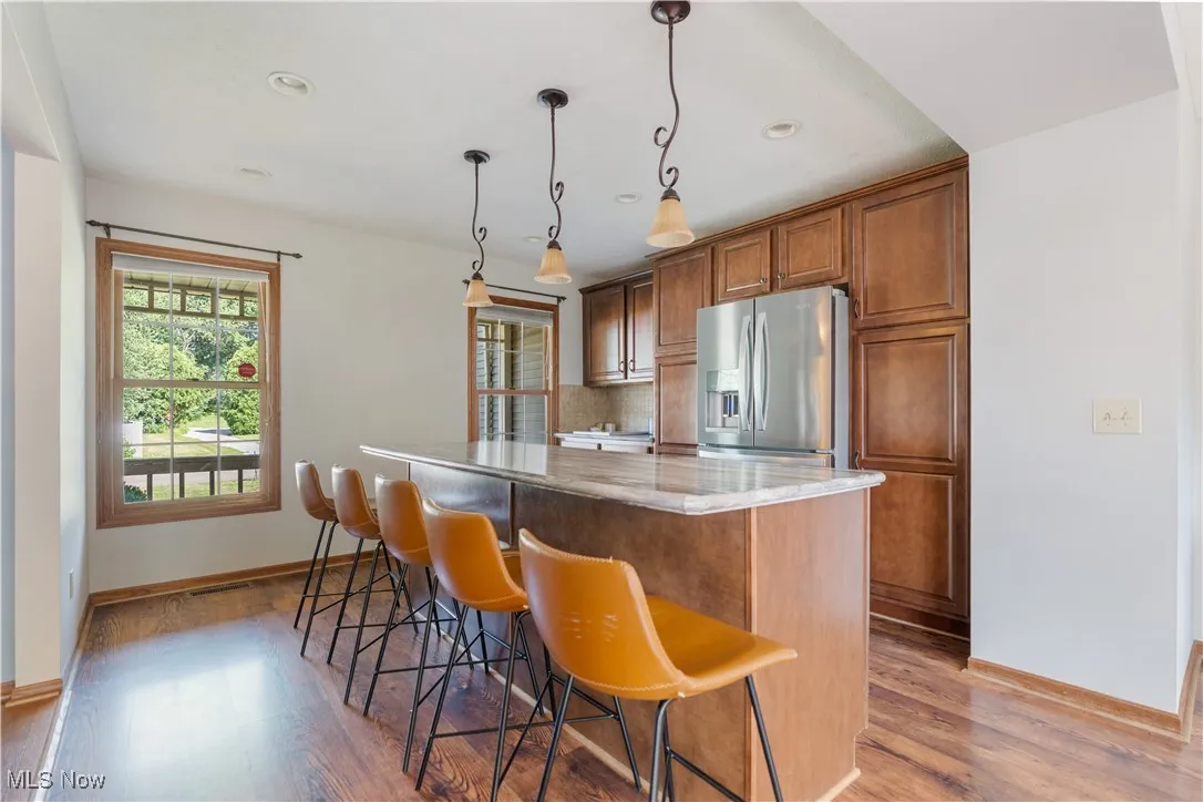 Kitchen with brown cabinetry, light stone counters, decorative light fixtures, stainless steel refrigerator with ice dispenser, and a breakfast bar area